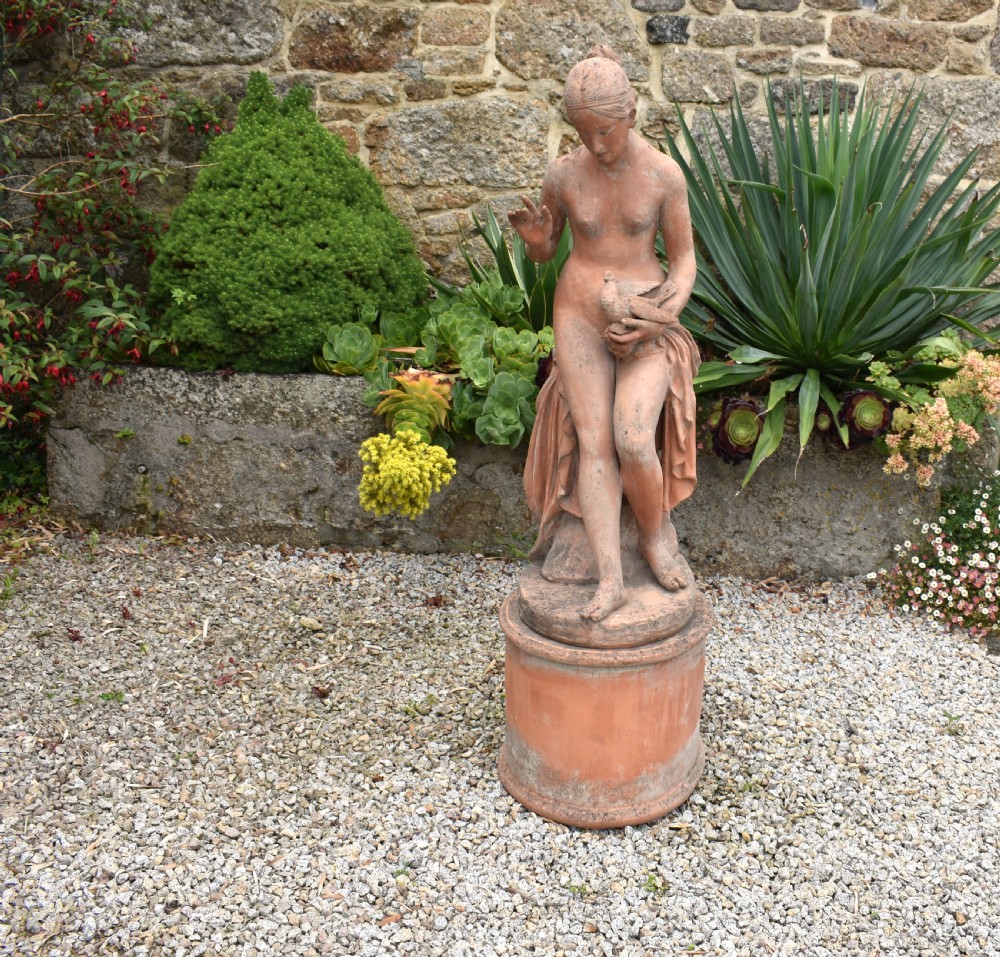 A classical terracotta sculpture of a seated draped female figure holding a bird, resting upon a matching oval terracotta pedestal. View III