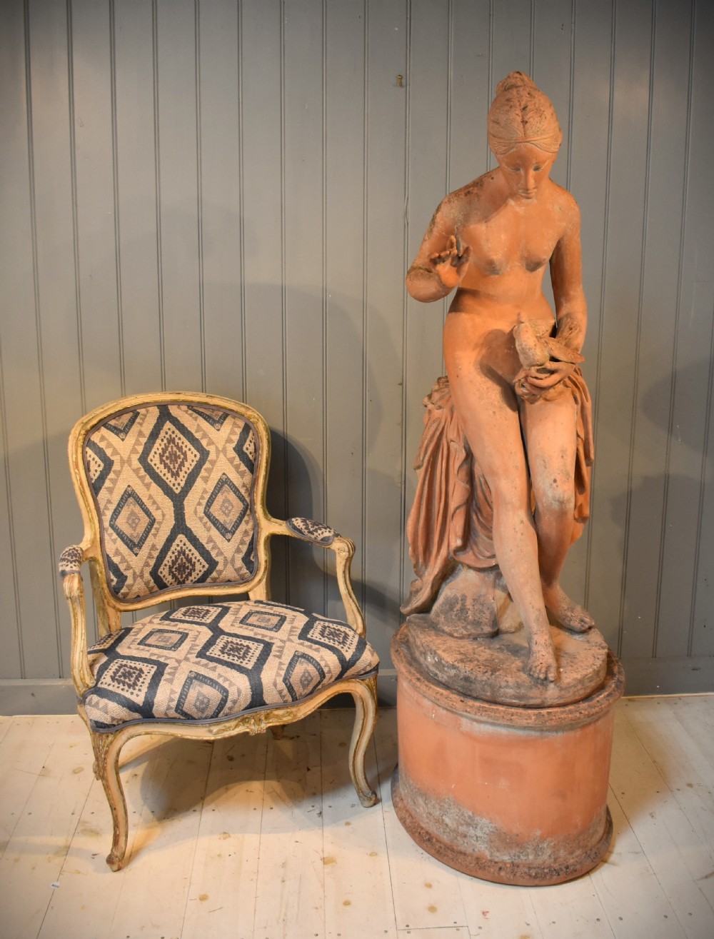 A classical terracotta sculpture of a seated draped female figure holding a bird, resting upon a matching oval terracotta pedestal. View IV