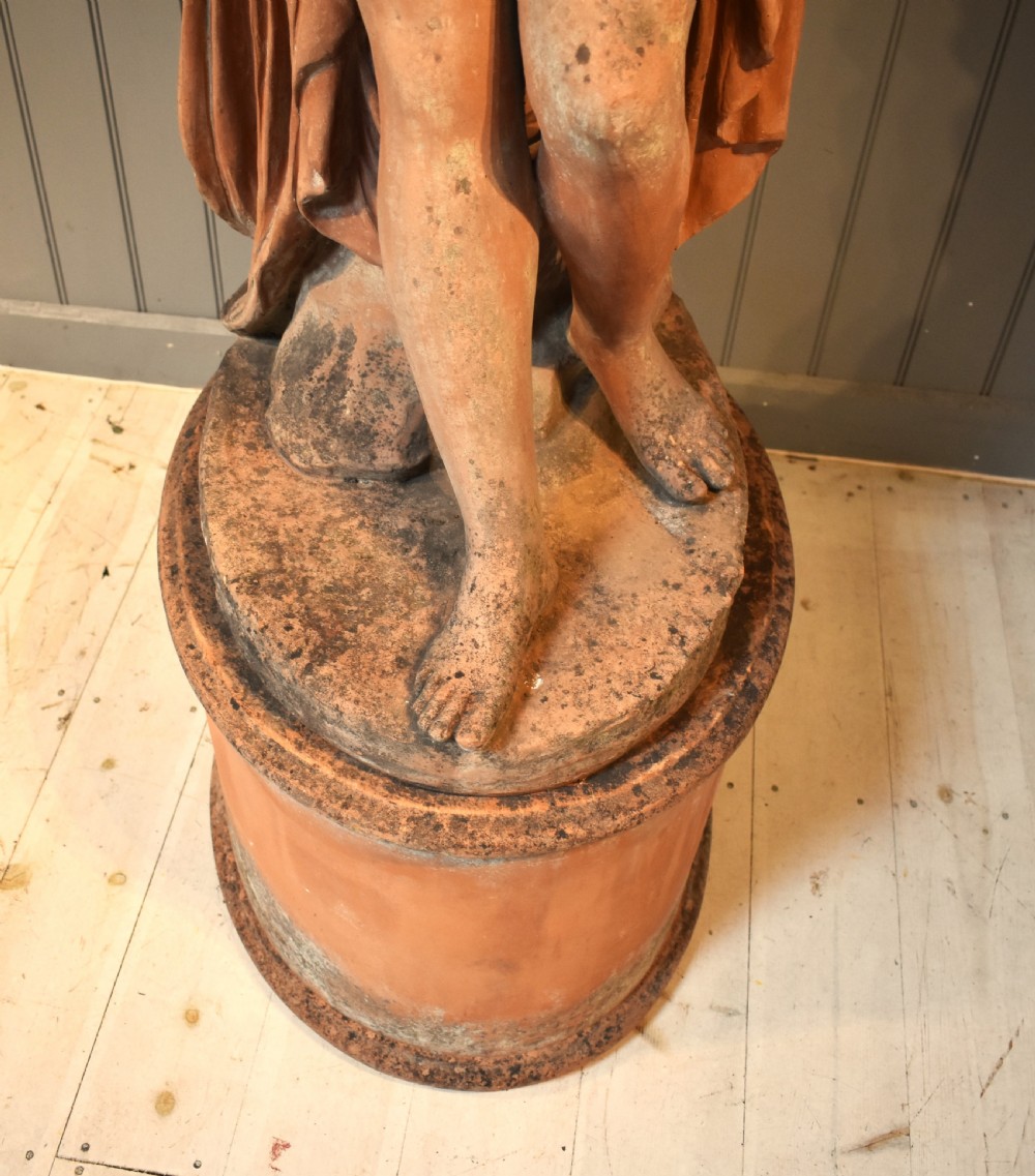 A classical terracotta sculpture of a seated draped female figure holding a bird, resting upon a matching oval terracotta pedestal. View VII