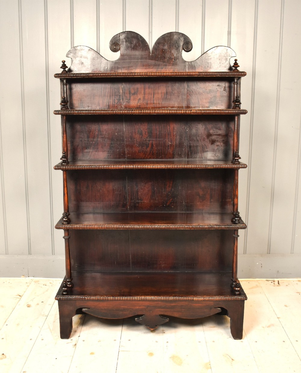 An antique dark mahogany open bookcase featuring four tiered shelves, turned spindle supports, beaded edging, and a carved scrolled crest.