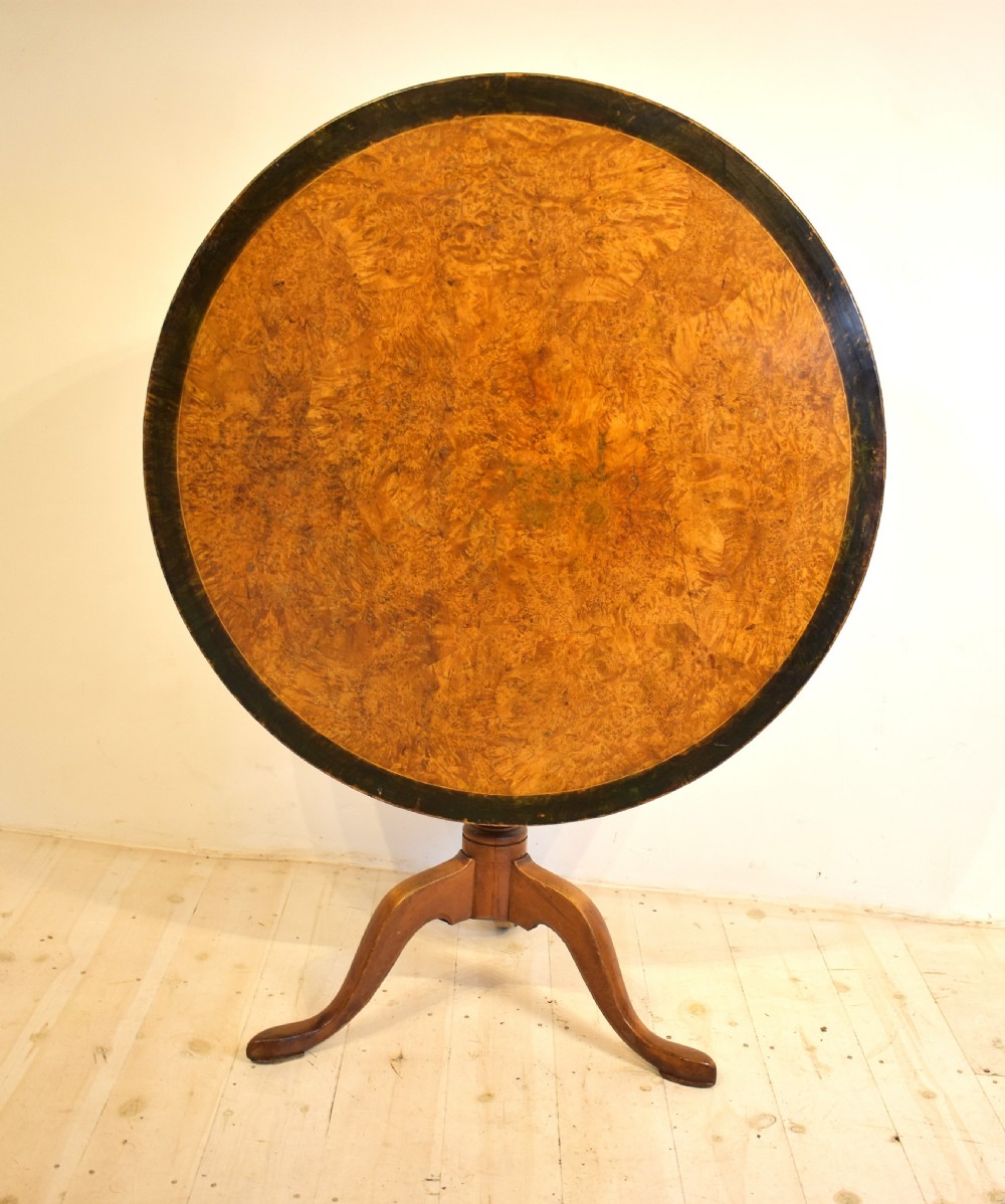 A circular burr wood tilt top table shown in its vertical position, supported by a turned central column and three cabriole legs.