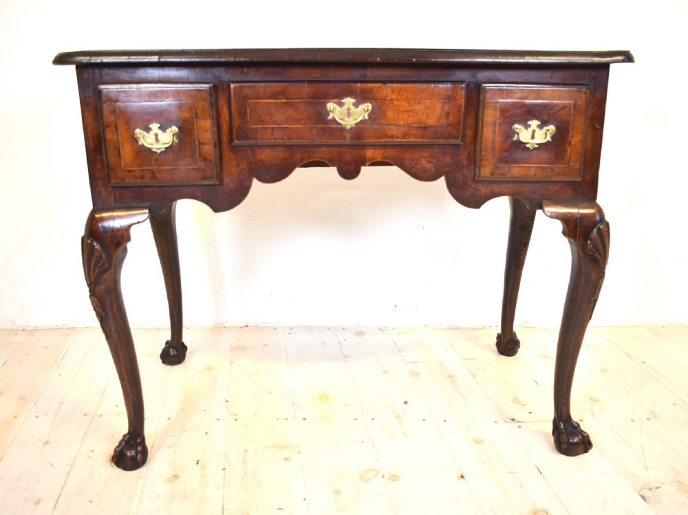 An antique Queen Anne walnut lowboy table featuring three drawers, brass handles, a shaped apron, and carved cabriole legs ending in ball and claw feet.