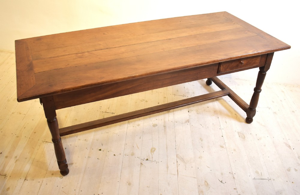 A rustic wooden farmhouse dining table featuring a plank top, a single side drawer, turned legs, and an H-stretcher base.