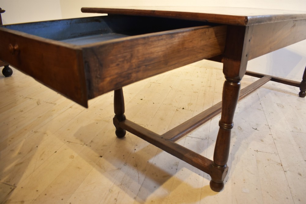 A rustic wooden farmhouse dining table featuring a plank top, a single side drawer, turned legs, and an H-stretcher base. View IV