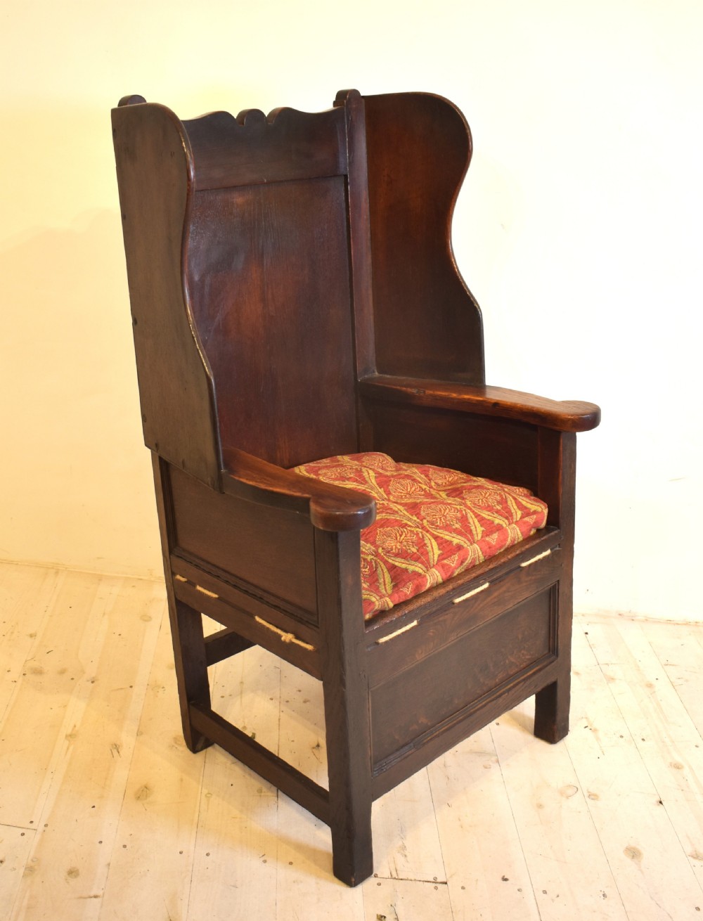 An antique dark oak lambing armchair with high winged sides, a panelled back, a storage base, and a patterned red seat cushion.
