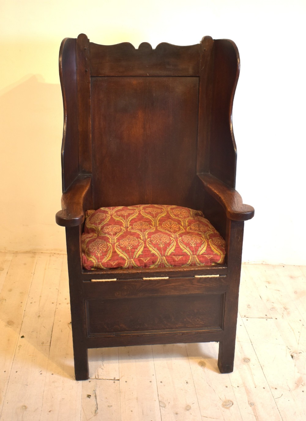 An antique dark oak lambing armchair with high winged sides, a panelled back, a storage base, and a patterned red seat cushion. View II