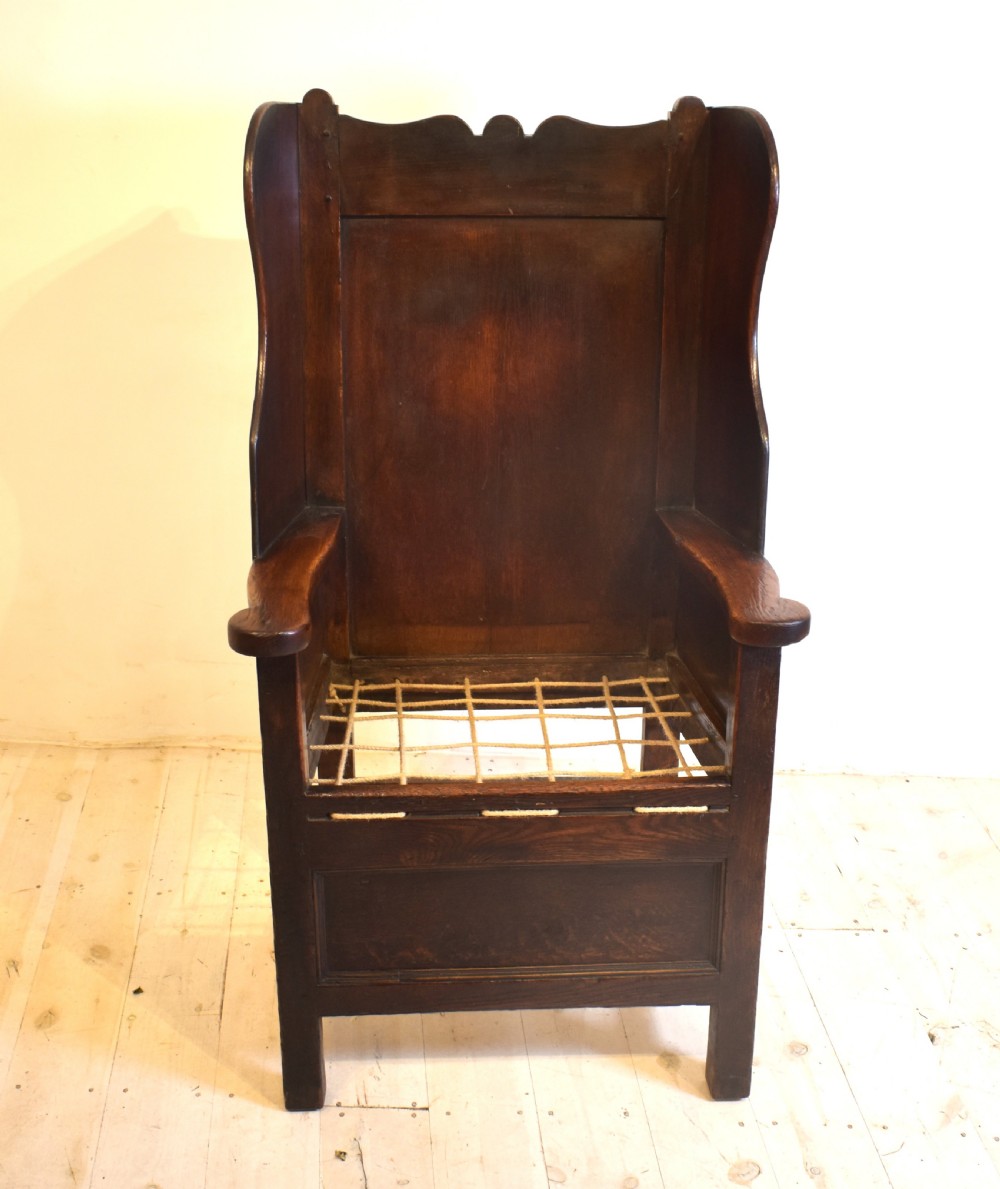 An antique dark oak lambing armchair with high winged sides, a panelled back, a storage base, and a patterned red seat cushion. View III