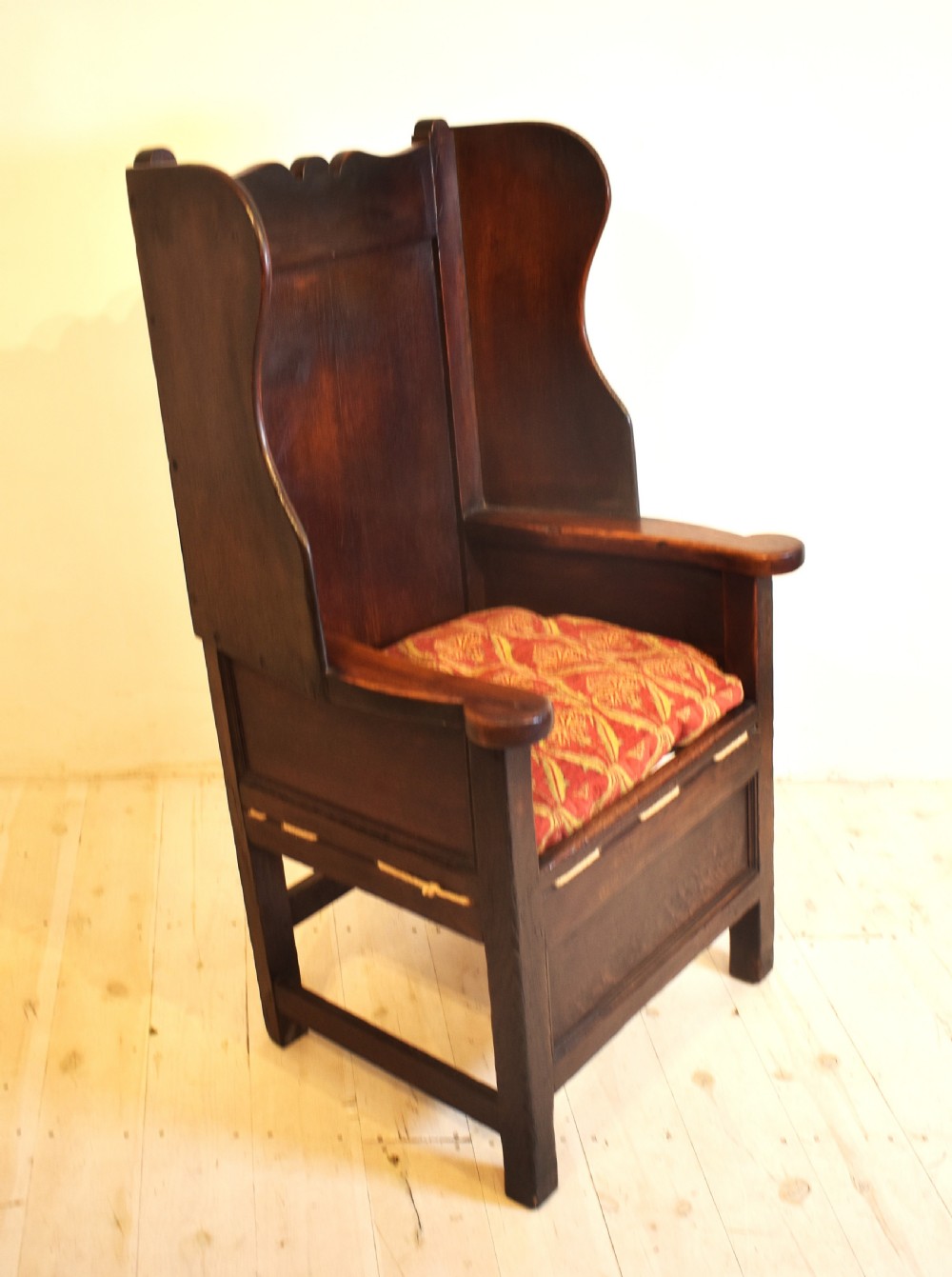 An antique dark oak lambing armchair with high winged sides, a panelled back, a storage base, and a patterned red seat cushion. View IV
