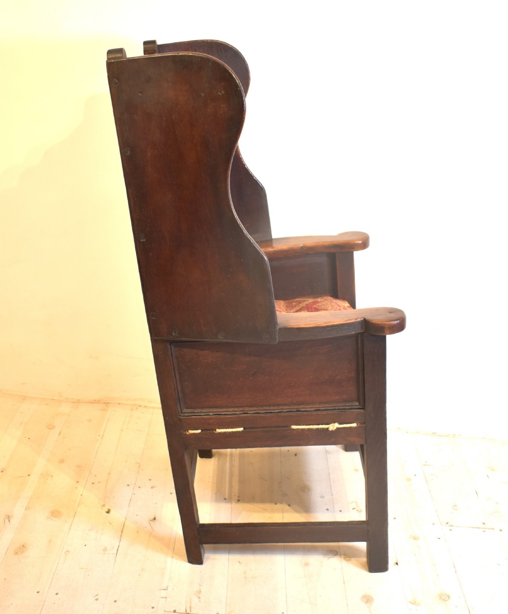 An antique dark oak lambing armchair with high winged sides, a panelled back, a storage base, and a patterned red seat cushion. View VI