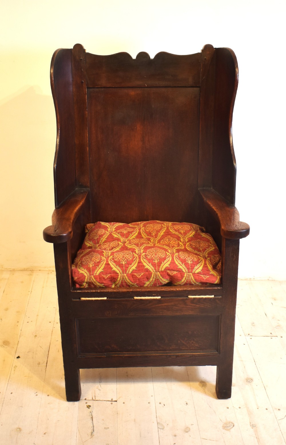An antique dark oak lambing armchair with high winged sides, a panelled back, a storage base, and a patterned red seat cushion. View VII