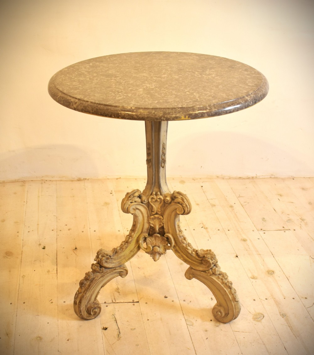A circular grey marble top table supported by a carved cream painted pedestal base with tripod scroll feet.