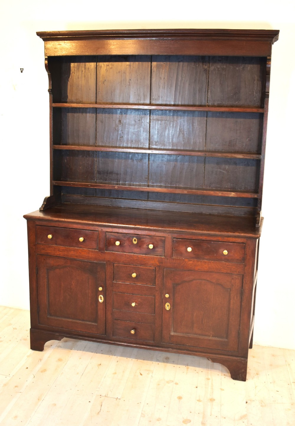 An antique dark oak Welsh dresser featuring a three-tier open plate rack above a base with multiple drawers, panelled cupboard doors, and brass knob handles.