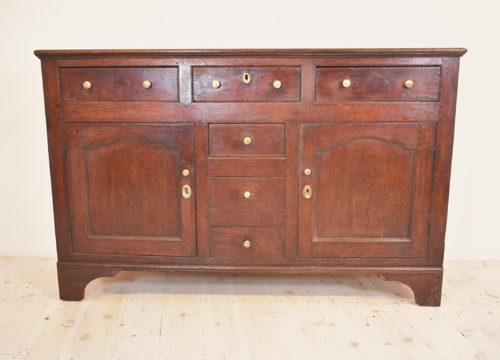An antique dark oak Welsh dresser featuring a three-tier open plate rack above a base with multiple drawers, panelled cupboard doors, and brass knob handles. View III