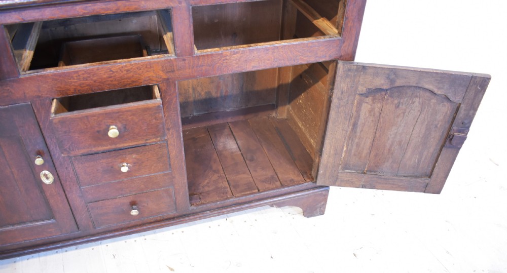 An antique dark oak Welsh dresser featuring a three-tier open plate rack above a base with multiple drawers, panelled cupboard doors, and brass knob handles. View VI