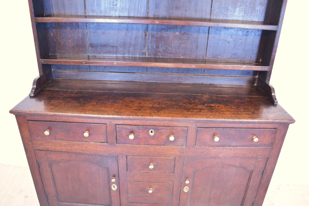 An antique dark oak Welsh dresser featuring a three-tier open plate rack above a base with multiple drawers, panelled cupboard doors, and brass knob handles. View VII