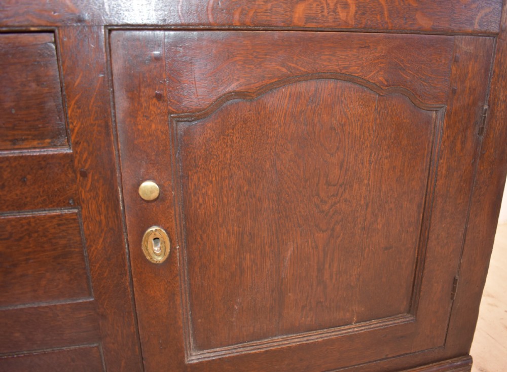 An antique dark oak Welsh dresser featuring a three-tier open plate rack above a base with multiple drawers, panelled cupboard doors, and brass knob handles. View IX