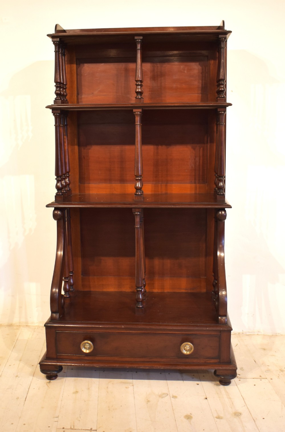 A dark mahogany waterfall bookcase featuring four graduated open shelves supported by turned spindles, curved side supports, and a base drawer with two brass knobs. View IV
