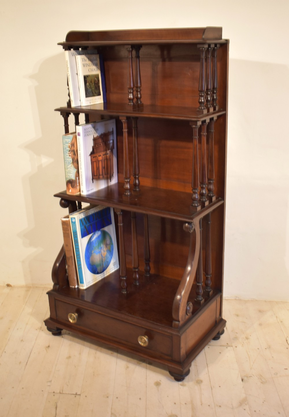 A dark mahogany waterfall bookcase featuring four graduated open shelves supported by turned spindles, curved side supports, and a base drawer with two brass knobs. View XI