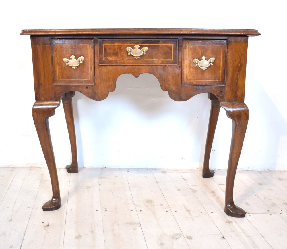 An antique Queen Anne style walnut lowboy featuring three crossbanded drawers with brass drop handles, a scalloped apron, and slender cabriole legs.