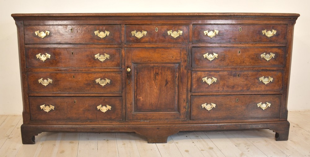 An eighteenth-century dark oak Lancashire dresser base featuring a central fielded panel cupboard door flanked by multiple drawers with brass batwing drop handles.