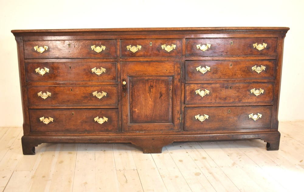 An eighteenth-century dark oak Lancashire dresser base featuring a central fielded panel cupboard door flanked by multiple drawers with brass batwing drop handles. View II