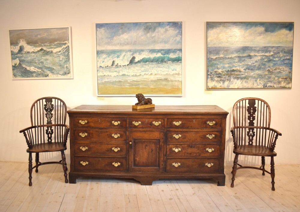 An eighteenth-century dark oak Lancashire dresser base featuring a central fielded panel cupboard door flanked by multiple drawers with brass batwing drop handles. View III