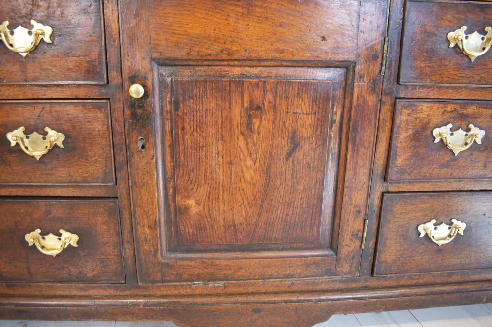 An eighteenth-century dark oak Lancashire dresser base featuring a central fielded panel cupboard door flanked by multiple drawers with brass batwing drop handles. View IV