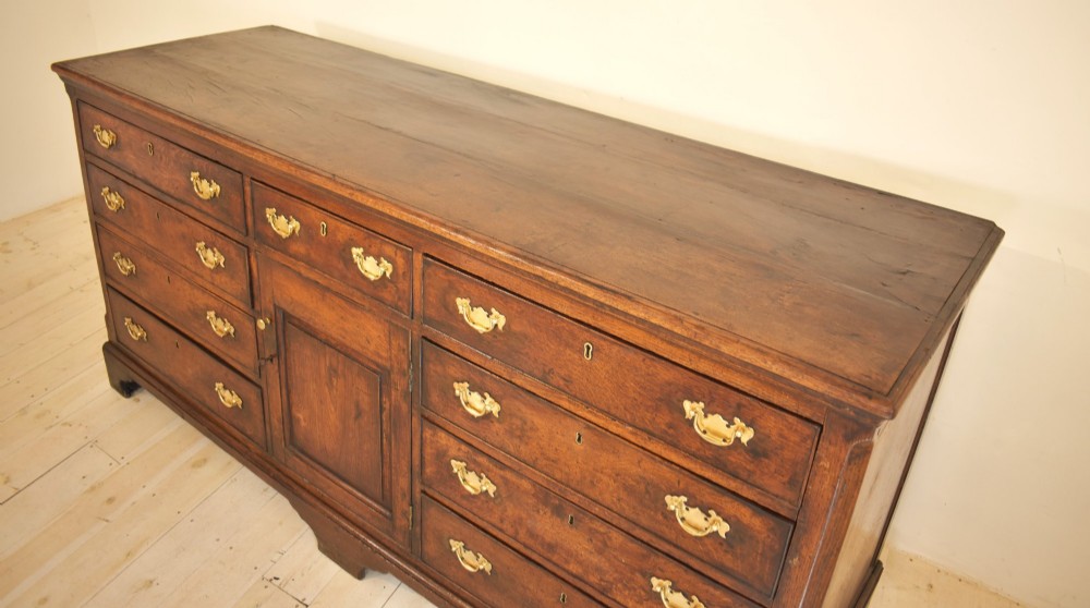 An eighteenth-century dark oak Lancashire dresser base featuring a central fielded panel cupboard door flanked by multiple drawers with brass batwing drop handles. View VI