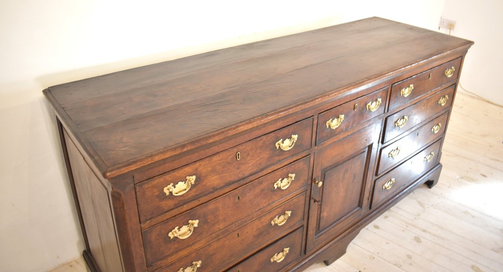 An eighteenth-century dark oak Lancashire dresser base featuring a central fielded panel cupboard door flanked by multiple drawers with brass batwing drop handles. View VII