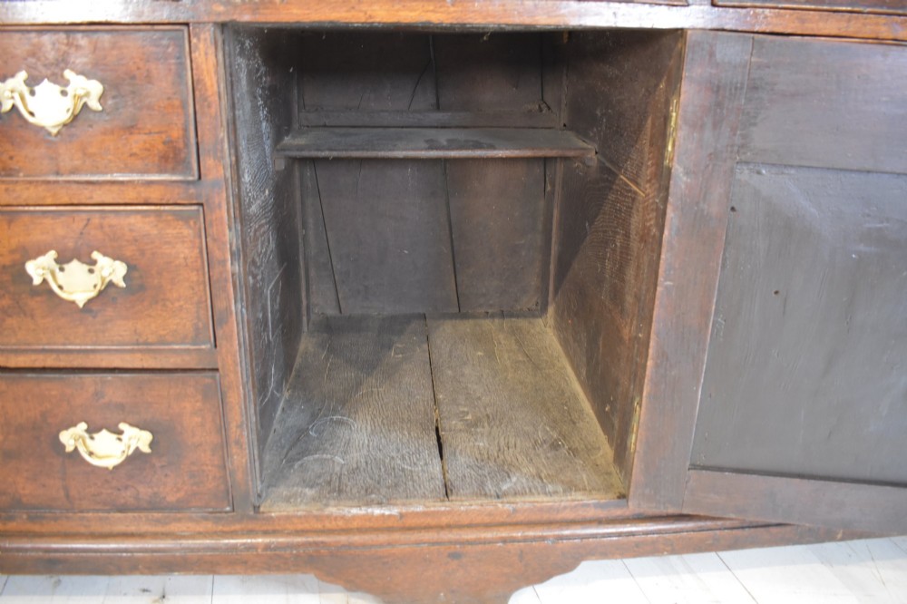 An eighteenth-century dark oak Lancashire dresser base featuring a central fielded panel cupboard door flanked by multiple drawers with brass batwing drop handles. View X