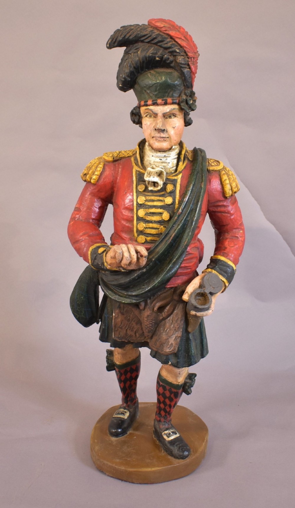 A carved and painted wooden figure of a Scottish Highlander soldier wearing a red jacket, kilt, and feathered bonnet.