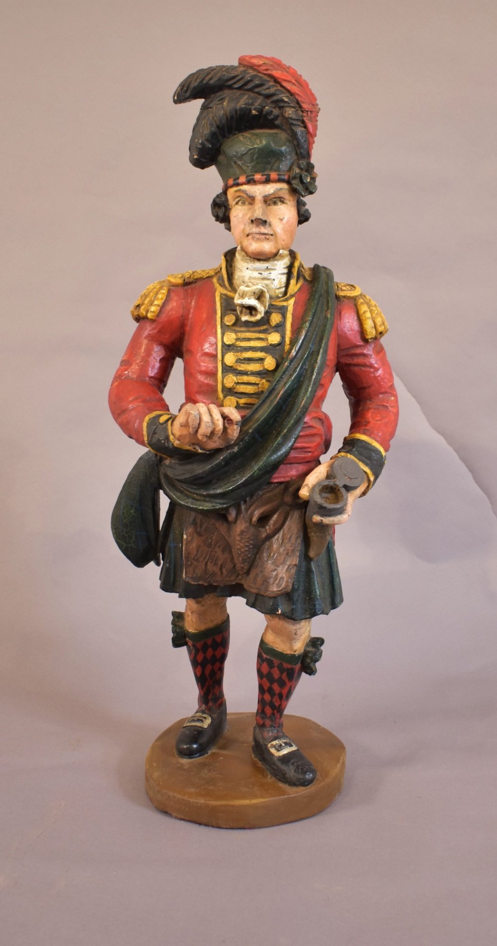 A carved and painted wooden figure of a Scottish Highlander soldier wearing a red jacket, kilt, and feathered bonnet. View II