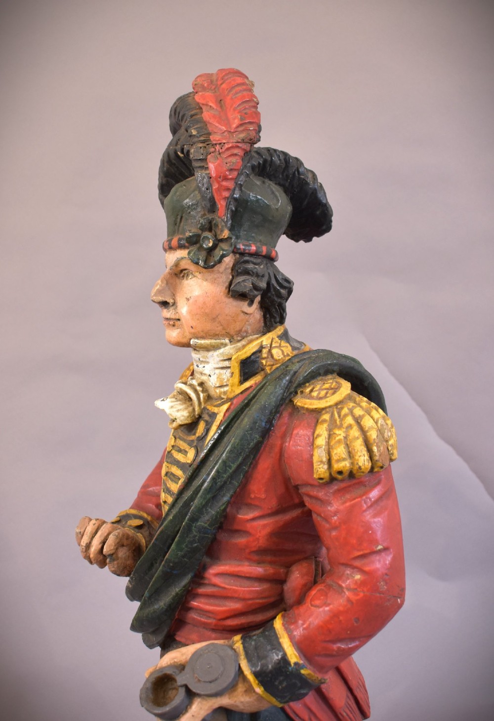 A carved and painted wooden figure of a Scottish Highlander soldier wearing a red jacket, kilt, and feathered bonnet. View III