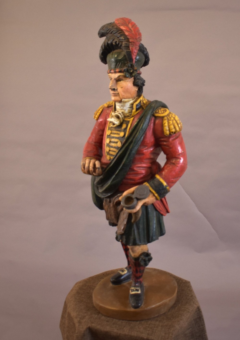 A carved and painted wooden figure of a Scottish Highlander soldier wearing a red jacket, kilt, and feathered bonnet. View IV