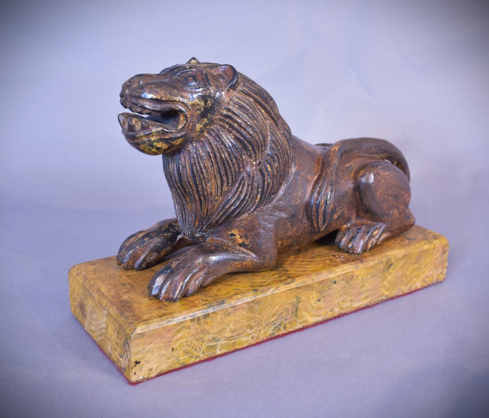 A hand-carved wooden recumbent lion figure featuring an open mouth and detailed mane, resting upon a rectangular gilded plinth.