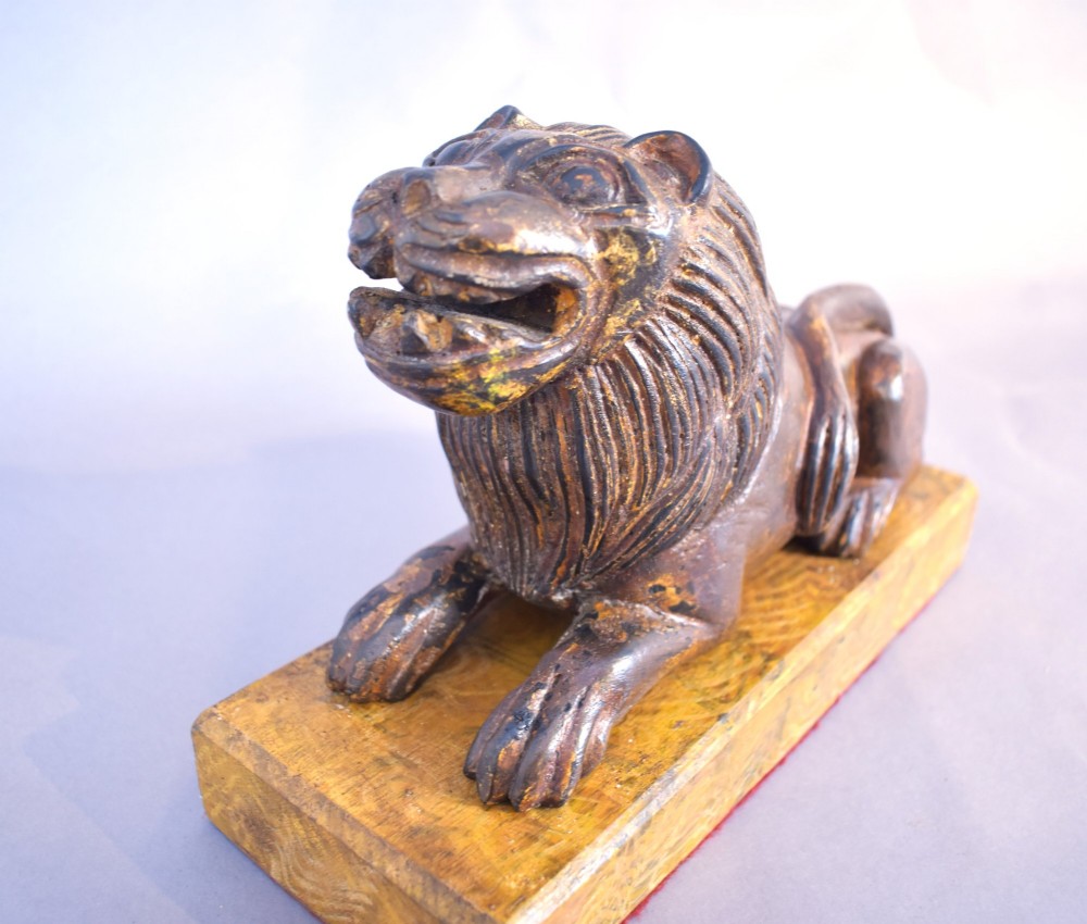 A hand-carved wooden recumbent lion figure featuring an open mouth and detailed mane, resting upon a rectangular gilded plinth. View II