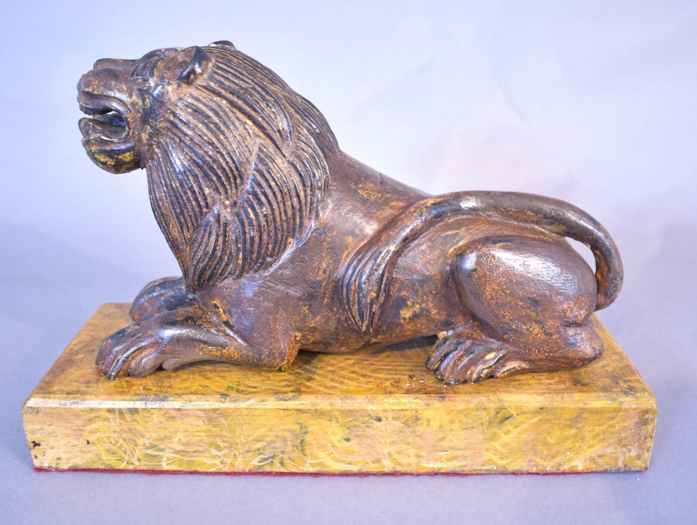A hand-carved wooden recumbent lion figure featuring an open mouth and detailed mane, resting upon a rectangular gilded plinth. View III