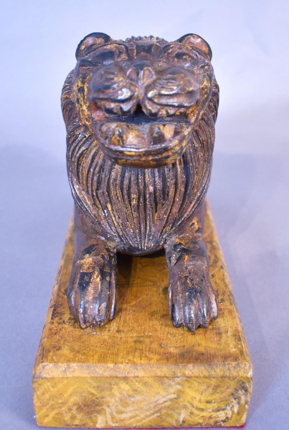 A hand-carved wooden recumbent lion figure featuring an open mouth and detailed mane, resting upon a rectangular gilded plinth. View IV