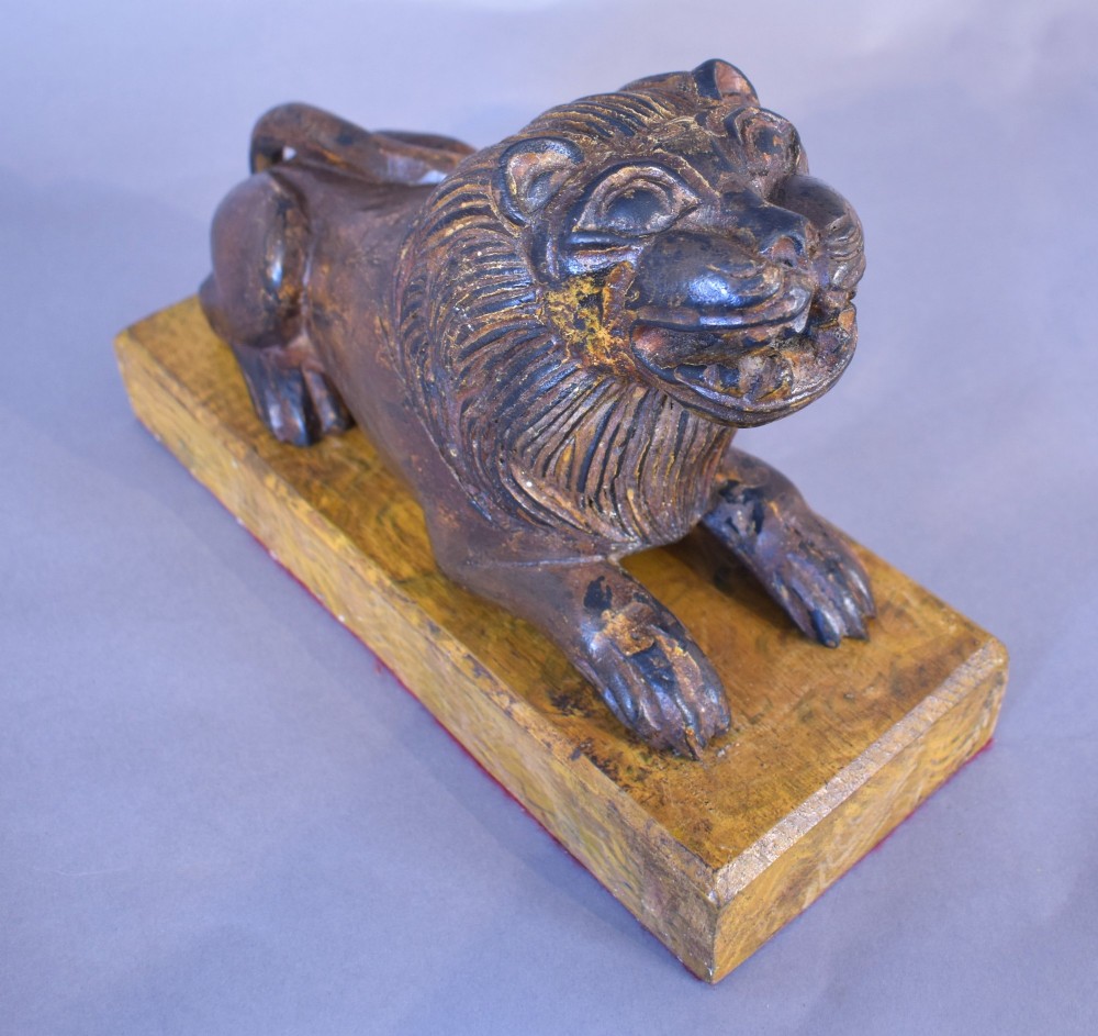 A hand-carved wooden recumbent lion figure featuring an open mouth and detailed mane, resting upon a rectangular gilded plinth. View V