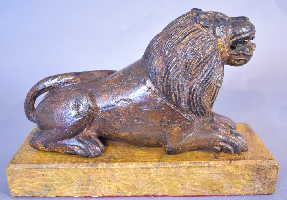 A hand-carved wooden recumbent lion figure featuring an open mouth and detailed mane, resting upon a rectangular gilded plinth. View VI