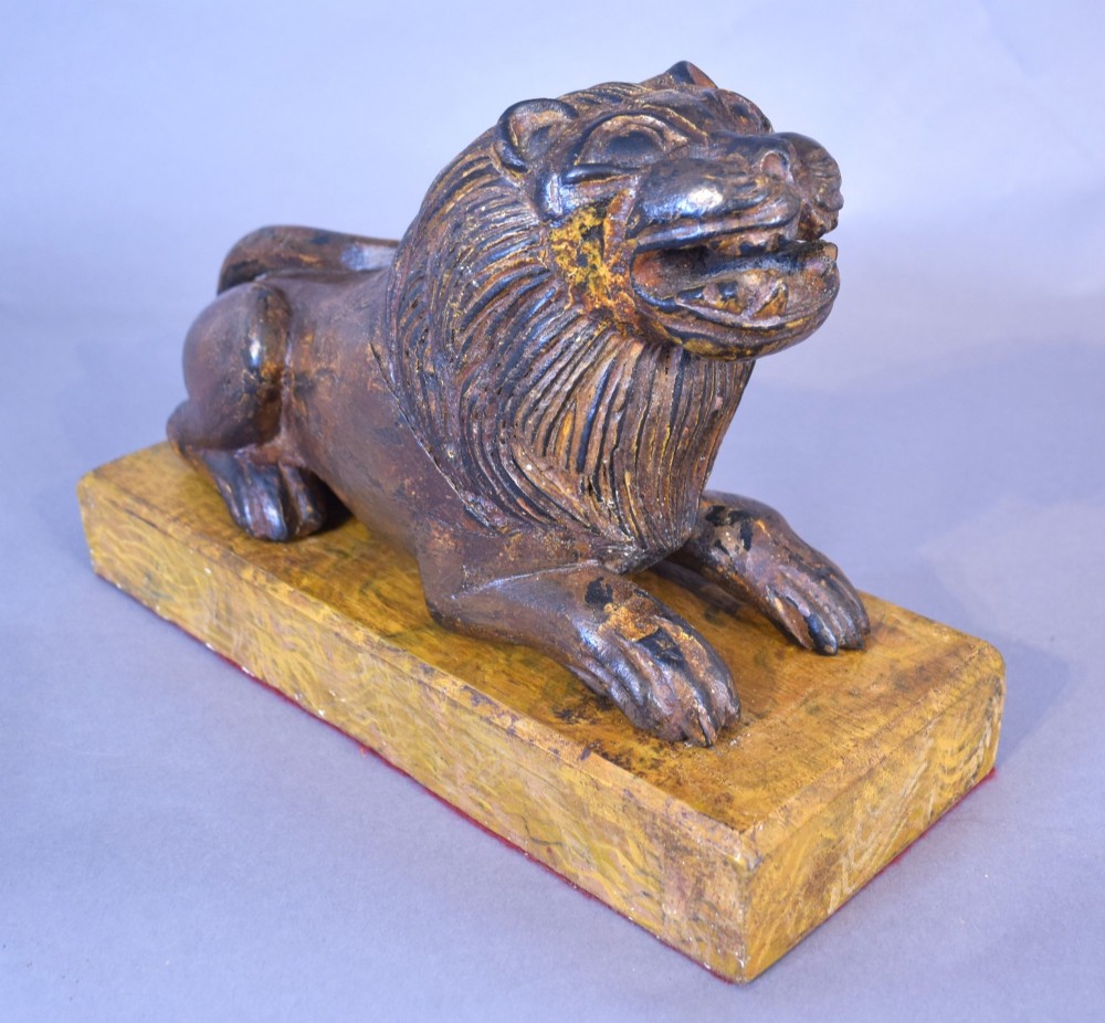 A hand-carved wooden recumbent lion figure featuring an open mouth and detailed mane, resting upon a rectangular gilded plinth. View VII