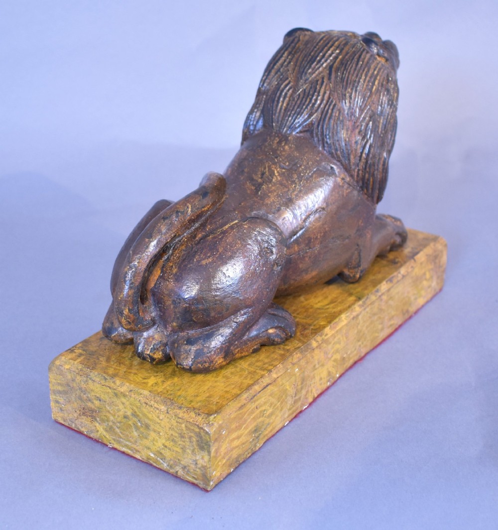 A hand-carved wooden recumbent lion figure featuring an open mouth and detailed mane, resting upon a rectangular gilded plinth. View VIII
