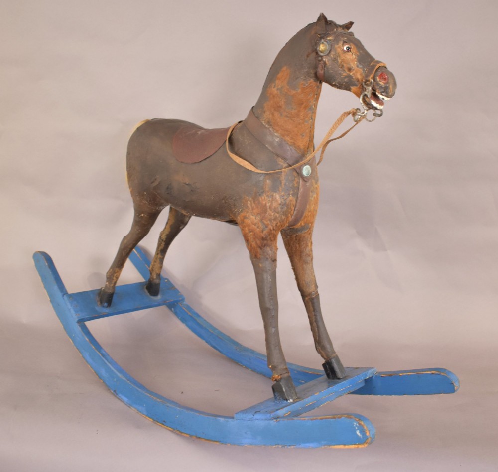 An antique wooden rocking horse with a dark distressed body, leather saddle, and yellowed hair tail, mounted on bright blue wooden bow rockers. View II