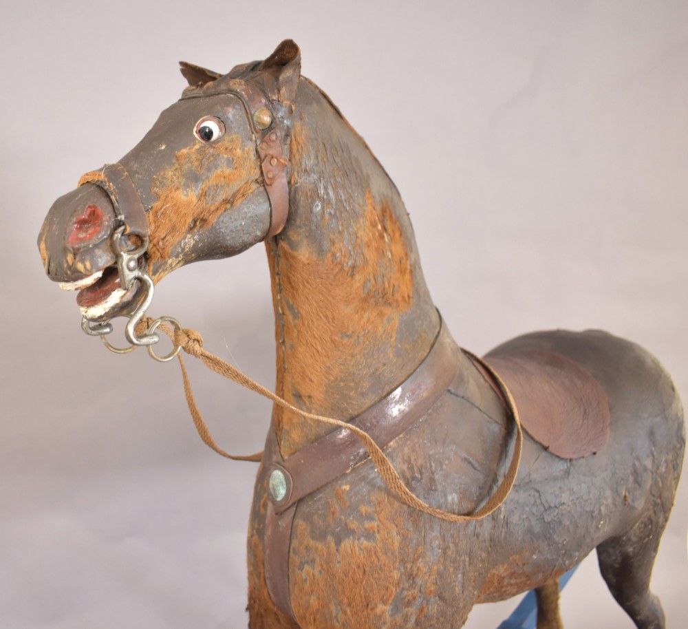 An antique wooden rocking horse with a dark distressed body, leather saddle, and yellowed hair tail, mounted on bright blue wooden bow rockers. View III