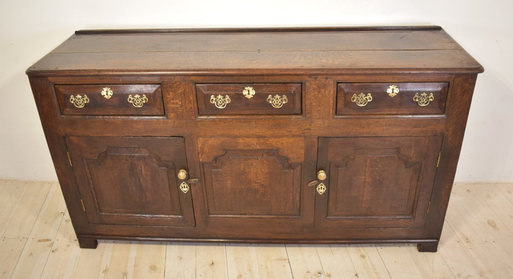 An 18th-century dark oak dresser base featuring three top drawers with brass handles above three cupboard doors with shaped fielded panels. View II
