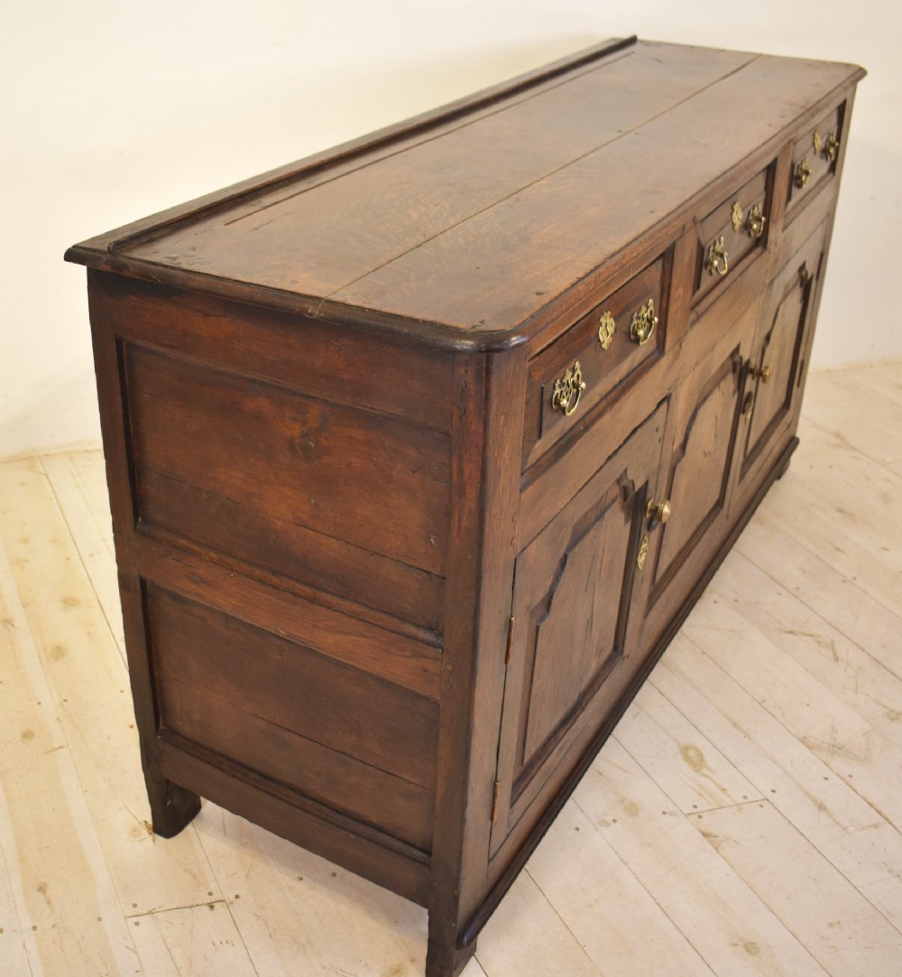 An 18th-century dark oak dresser base featuring three top drawers with brass handles above three cupboard doors with shaped fielded panels. View V