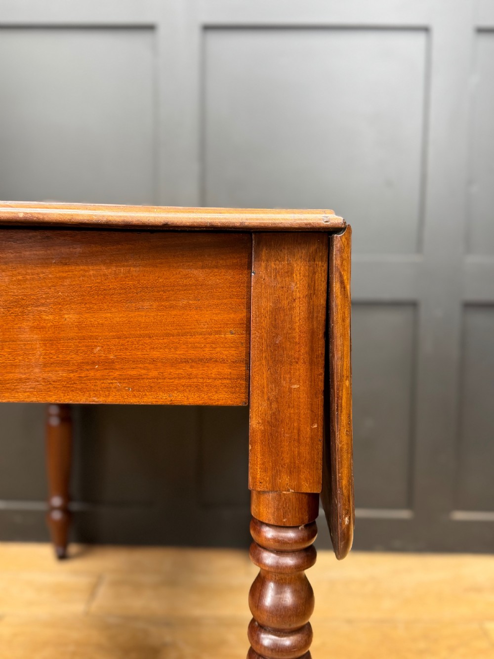 An antique mahogany drop leaf table with two raised side flaps and four turned legs set against a dark panelled backdrop. View V