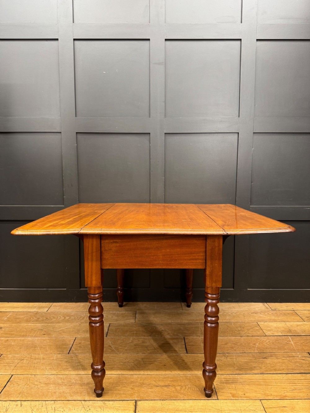 An antique mahogany drop leaf table with two raised side flaps and four turned legs set against a dark panelled backdrop.