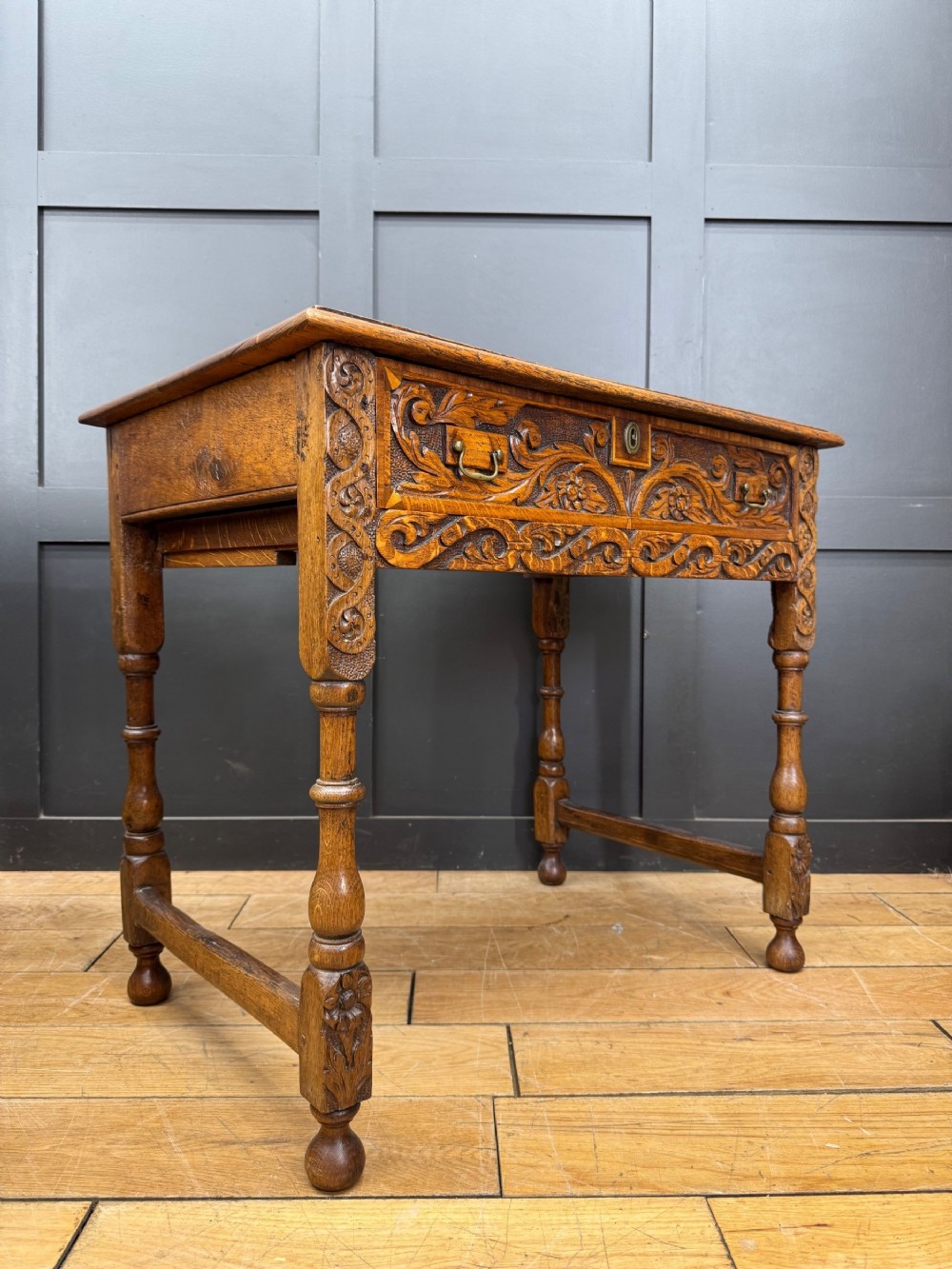 An antique carved oak side table featuring turned legs, a single carved drawer with brass handles, and lower stretchers.