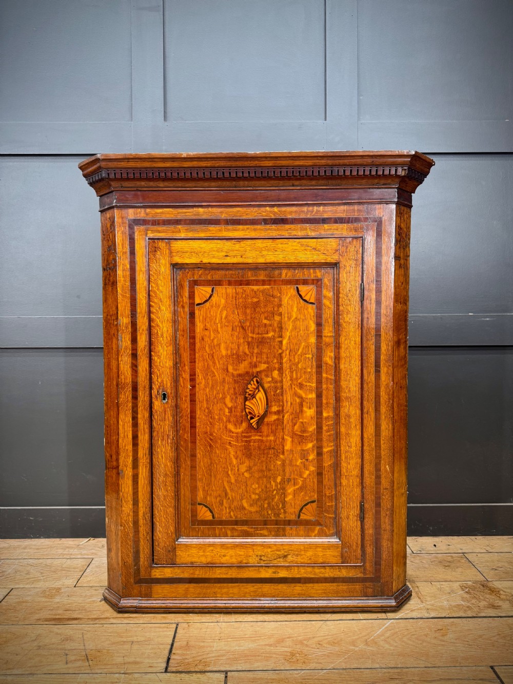 A Georgian oak wall corner cupboard featuring a single panel door with central shell marquetry inlay, crossbanded borders, and dentil moulding along the top cornice.